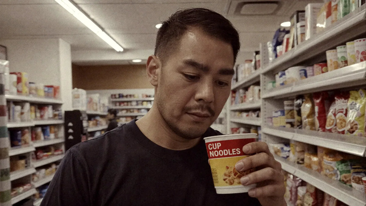 Alain in a convenience store aisle, looking at a cup of instant ramen