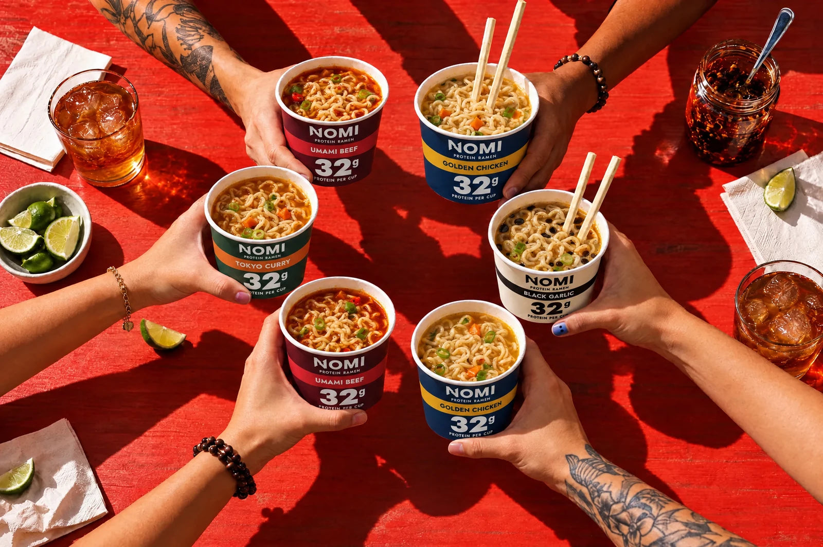 Six hands raising NOMI cups together over a red table